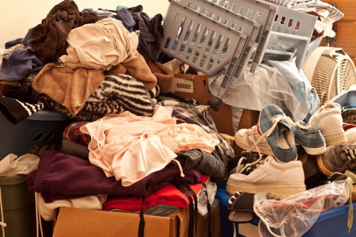 A large assortment of discarded clothing, textiles, and fabric items piled on top of each other, including sweaters, shirts, and trousers in various colours such as beige, red, maroon, and black. Some clothing appears crumpled, while others are folded or draped over the pile. Among the textiles, there are plastic laundry baskets, one grey and one beige, positioned at different angles, with some clothes spilling out of them. To the right, a pair of cream-coloured sneakers with blue accents is visible, partially covered by the pile. The scene is set indoors against a background of a wooden surface, possibly a floor or table, and the lighting is warm and even, emphasizing the variety of textures and colours. The image reflects a typical cluttered scene associated with rubbish or decluttering services offered by companies such as Office Clearance Chelsea, operating near Chelsea and the SW10 postcode area.