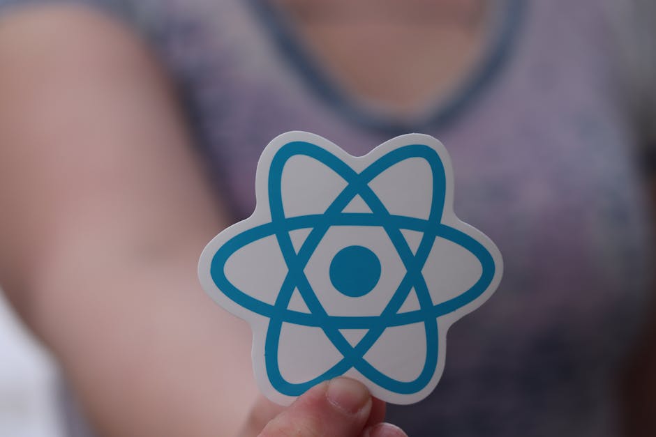A person's hand holding a white sticker with a blue atomic symbol design comprising intersecting elliptical orbits around a central circle. The background shows part of the person's upper arm and a bl